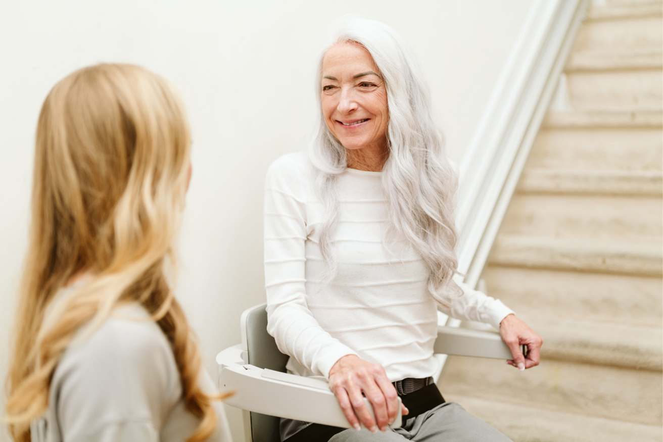 An elderly woman with long white hair sits on a stair lift, smiling at a younger woman with long blonde hair who is standing nearby.