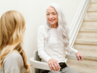 An elderly woman with long white hair sits on a stair lift, smiling at a younger woman with long blonde hair who is standing nearby.