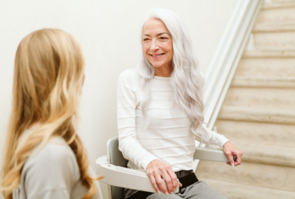 An elderly woman with long white hair sits on a stair lift, smiling at a younger woman with long blonde hair who is standing nearby.