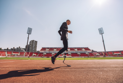 A man with a prosthetic leg runs on a track in an outdoor stadium under a clear sky.