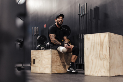 A man with a prosthetic leg sits on a wooden box in a gym, smiling and resting. Fitness equipment and weights are in the background.