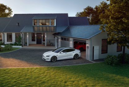 A modern house with solar panels on the roof, featuring a driveway with a white electric car parked in front. The garage door is open, and another car is partially visible inside.