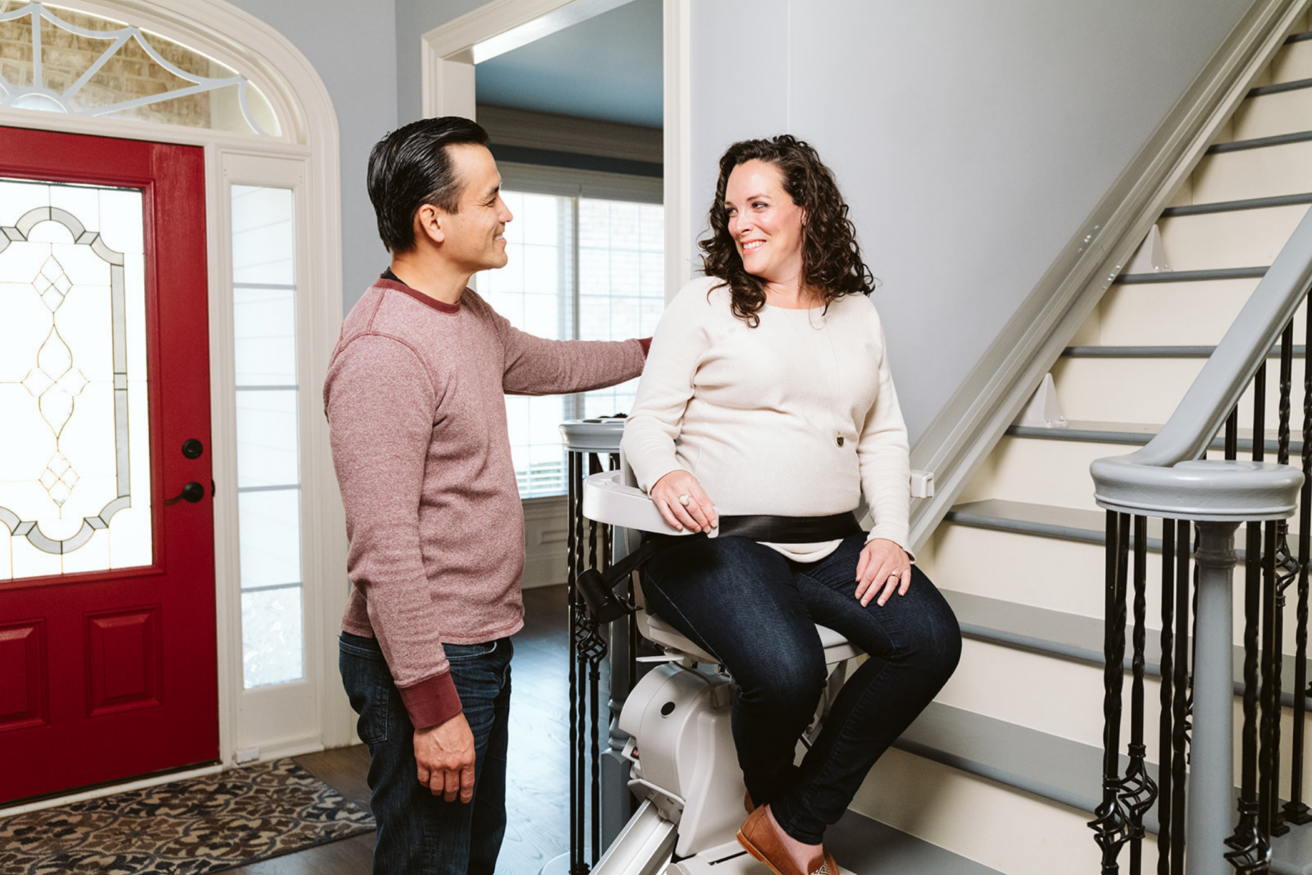 A man stands next to a woman seated on a stairlift at the bottom of a staircase inside a house. The woman is smiling, and both appear to be engaged in conversation.