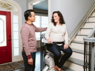 A man stands next to a woman seated on a stairlift at the bottom of a staircase inside a house. The woman is smiling, and both appear to be engaged in conversation.