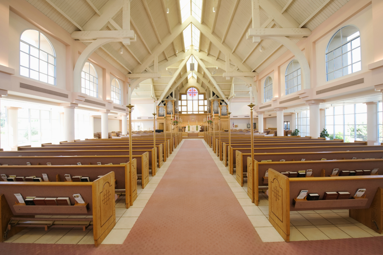 Interior of an empty church with wooden pews, a red carpet aisle, high vaulted ceiling, large windows, and a prominently displayed altar.