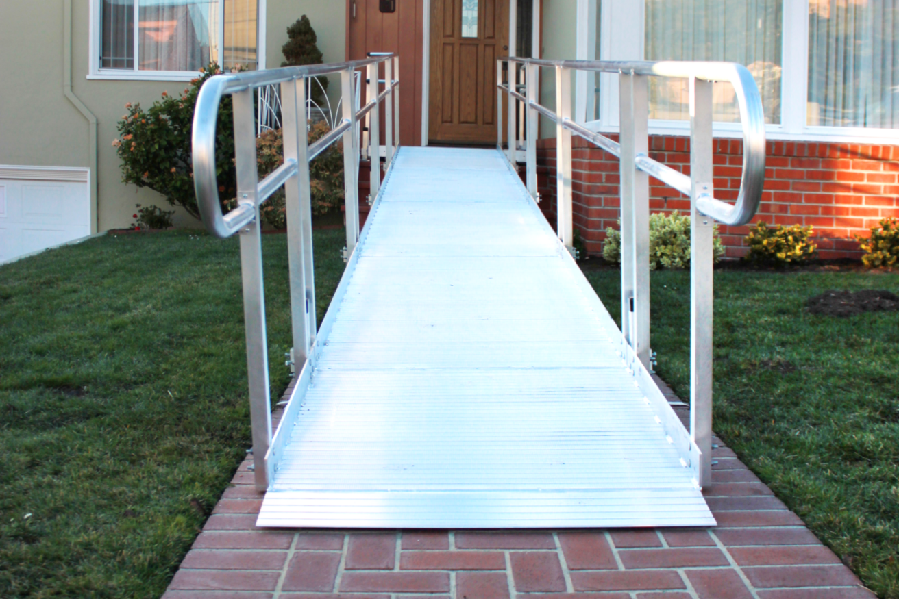 Aluminum wheelchair ramp leading to a building entrance, set over a brick walkway and surrounded by green grass.