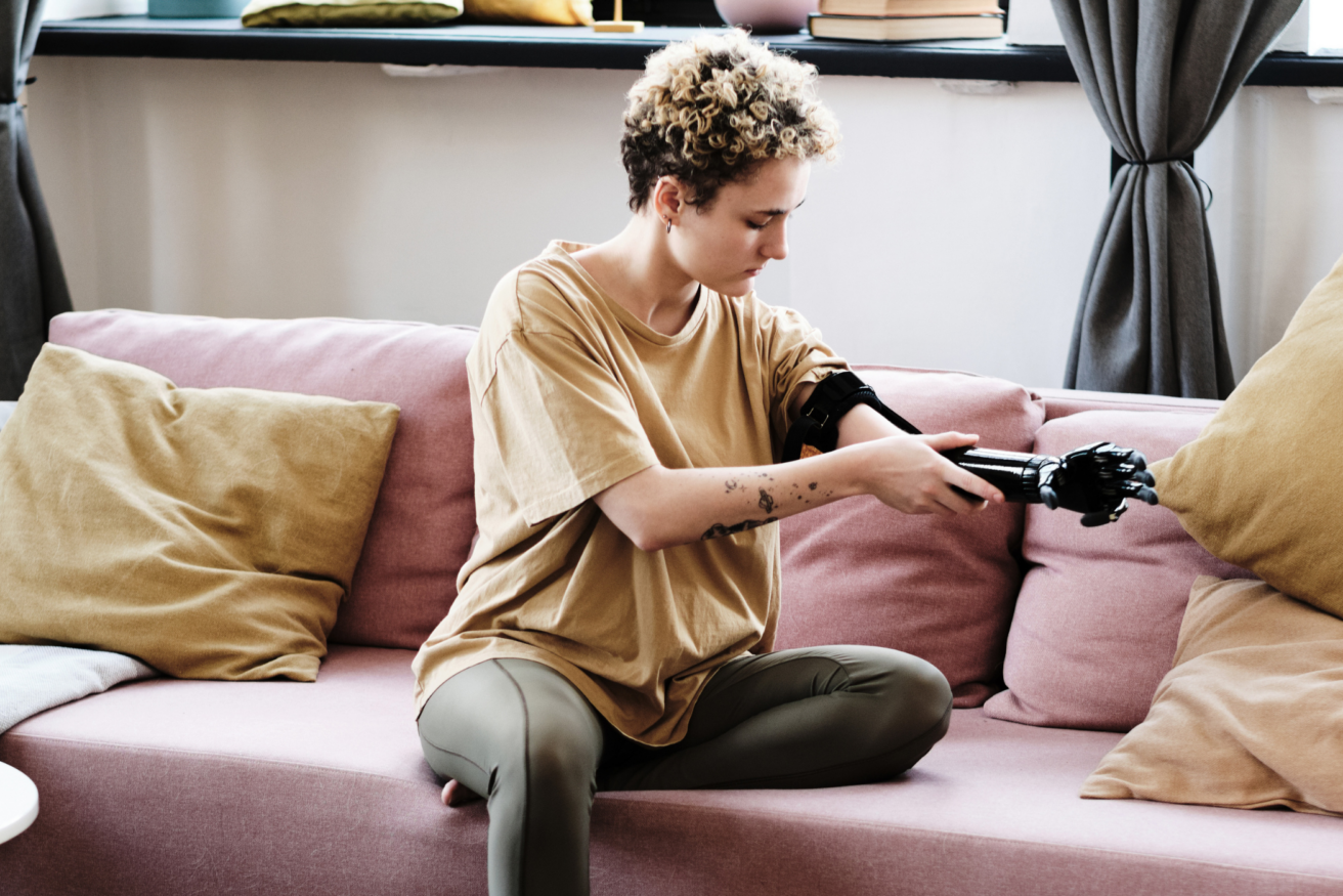 A person with short curly hair sits on a pink couch, adjusting their prosthetic arm with focused attention.