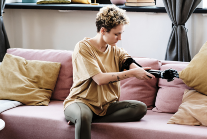 A person with short curly hair sits on a pink couch, adjusting their prosthetic arm with focused attention.