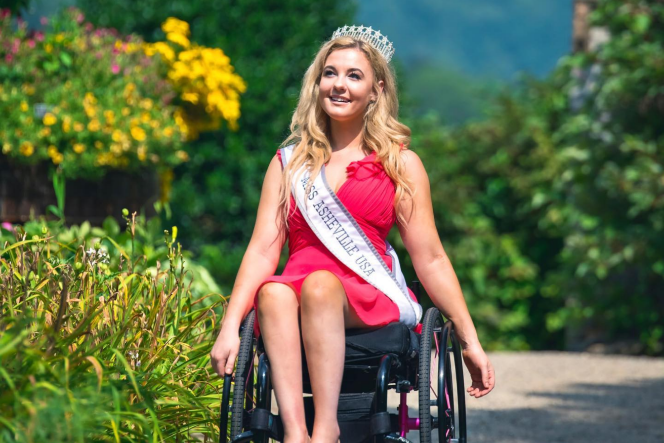 A woman wearing a crown and sash labeled "Miss Ashville USA" sits in a wheelchair, dressed in a red dress, with a garden background.