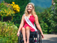 A woman wearing a crown and sash labeled "Miss Ashville USA" sits in a wheelchair, dressed in a red dress, with a garden background.