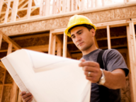 A construction worker wearing a yellow hard hat and gray shirt looks at blueprints inside a wooden building frame.