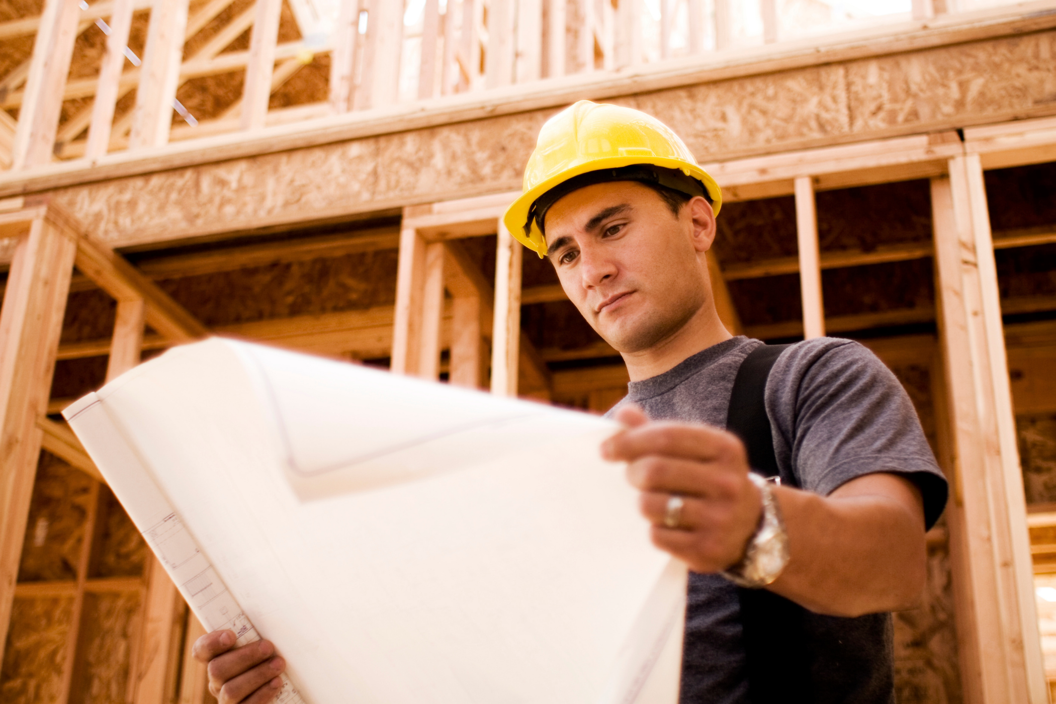 A construction worker wearing a yellow hard hat and gray shirt looks at blueprints inside a wooden building frame.