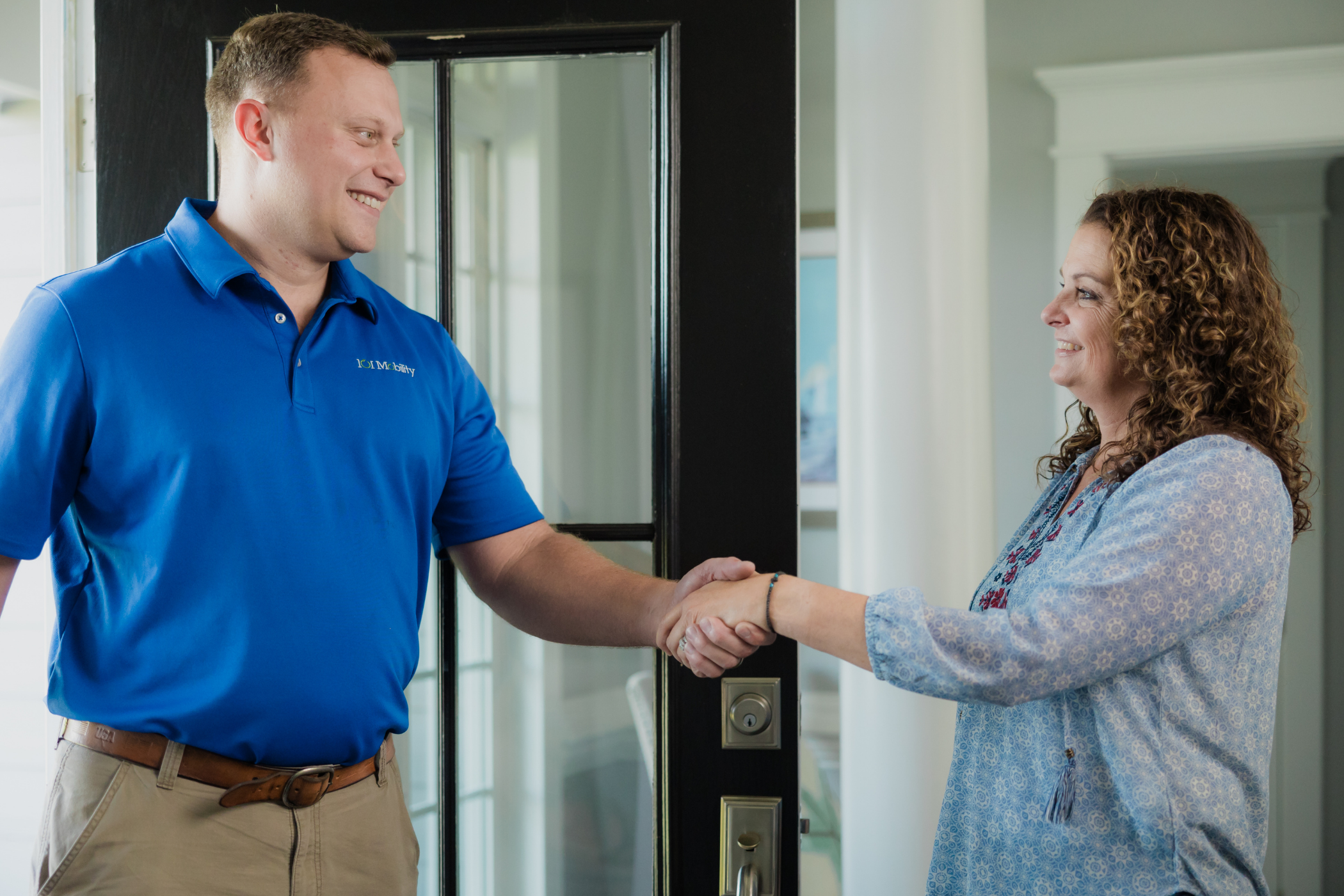 Two people shake hands at the doorway, one wearing a blue polo shirt and the other in a patterned blouse. The setting appears to be indoors with a glass door and white walls in the background.