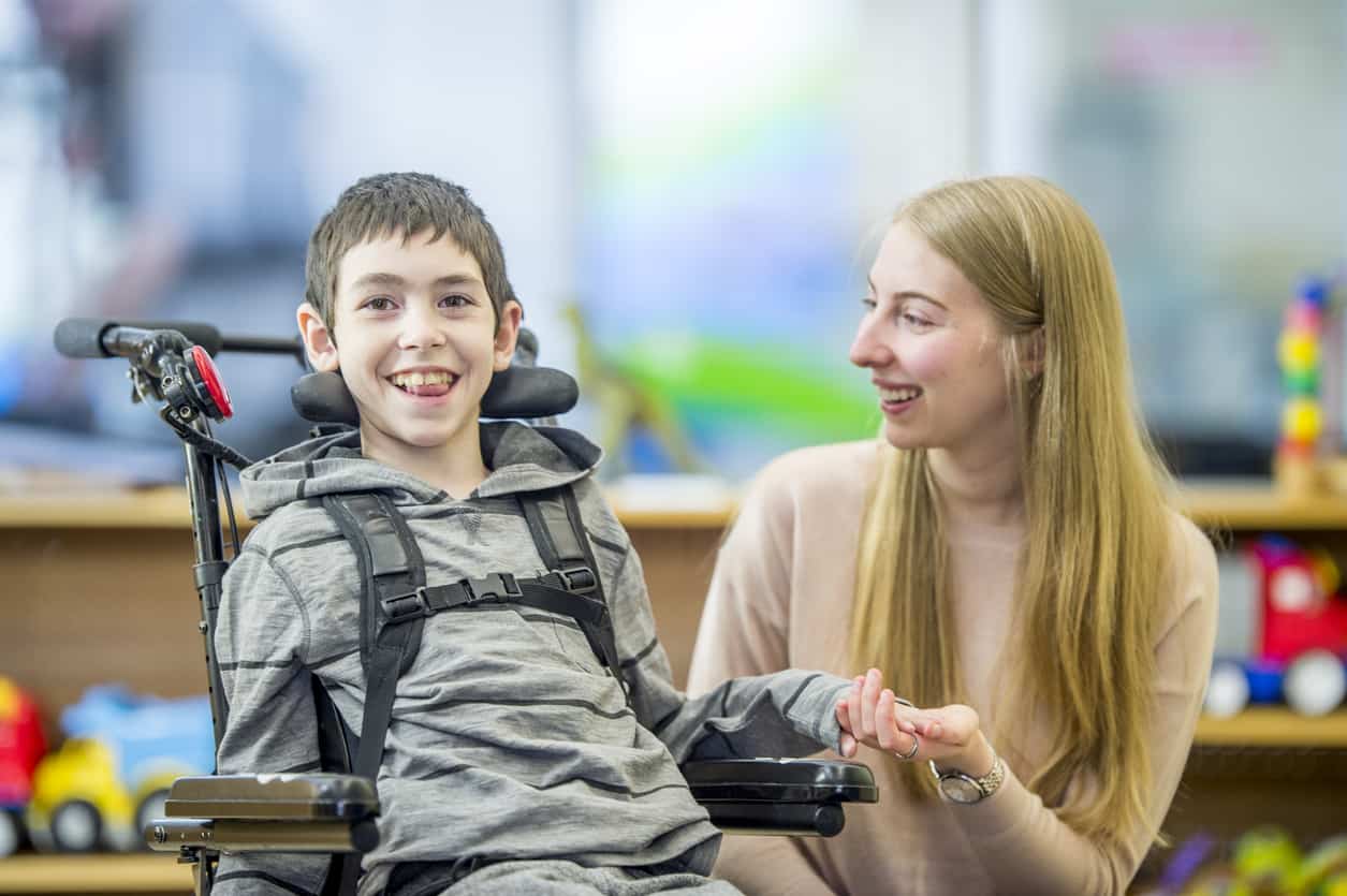 Little boy in wheelchair holds woman’s hand in classroom