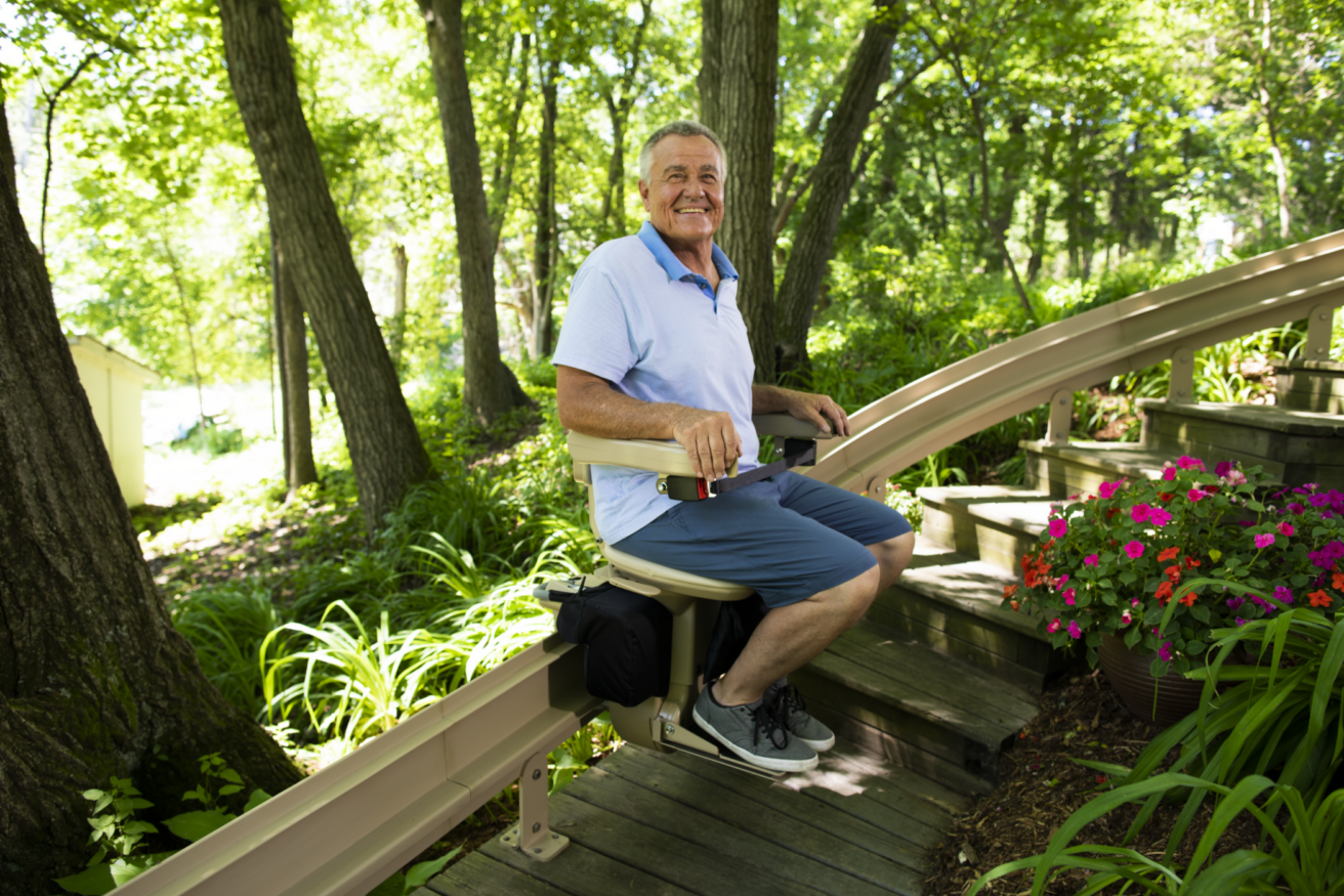 A senior man sits on an outdoor stairlift installed along a wooden staircase in a lush, green garden with colorful flowers.