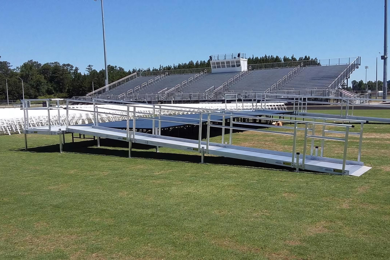 A large, empty stadium with metal bleachers and a metal ramp leading to the field, surrounded by green grass and clear skies.