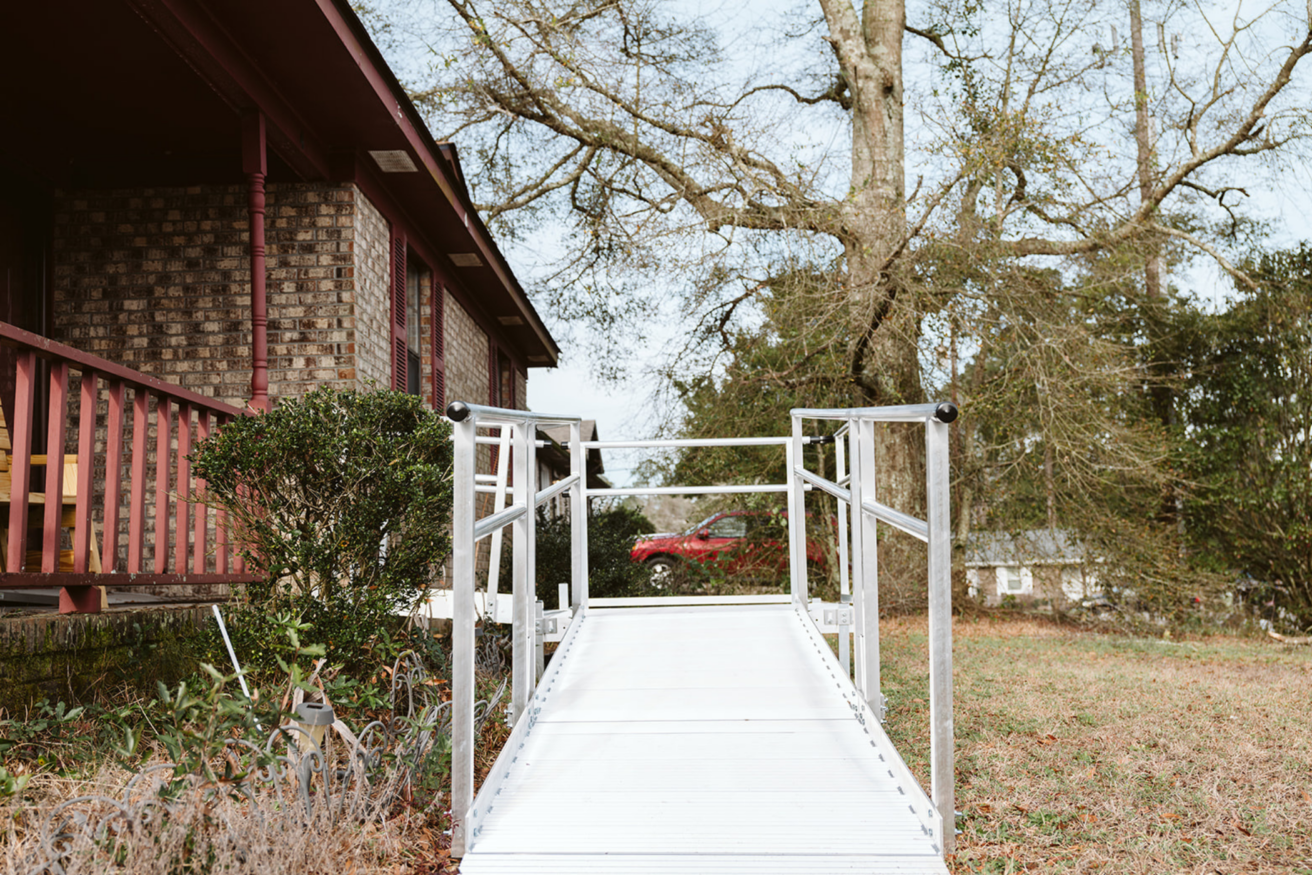 A metal ramp leads up to the porch of a brick house surrounded by trees and a grassy area.