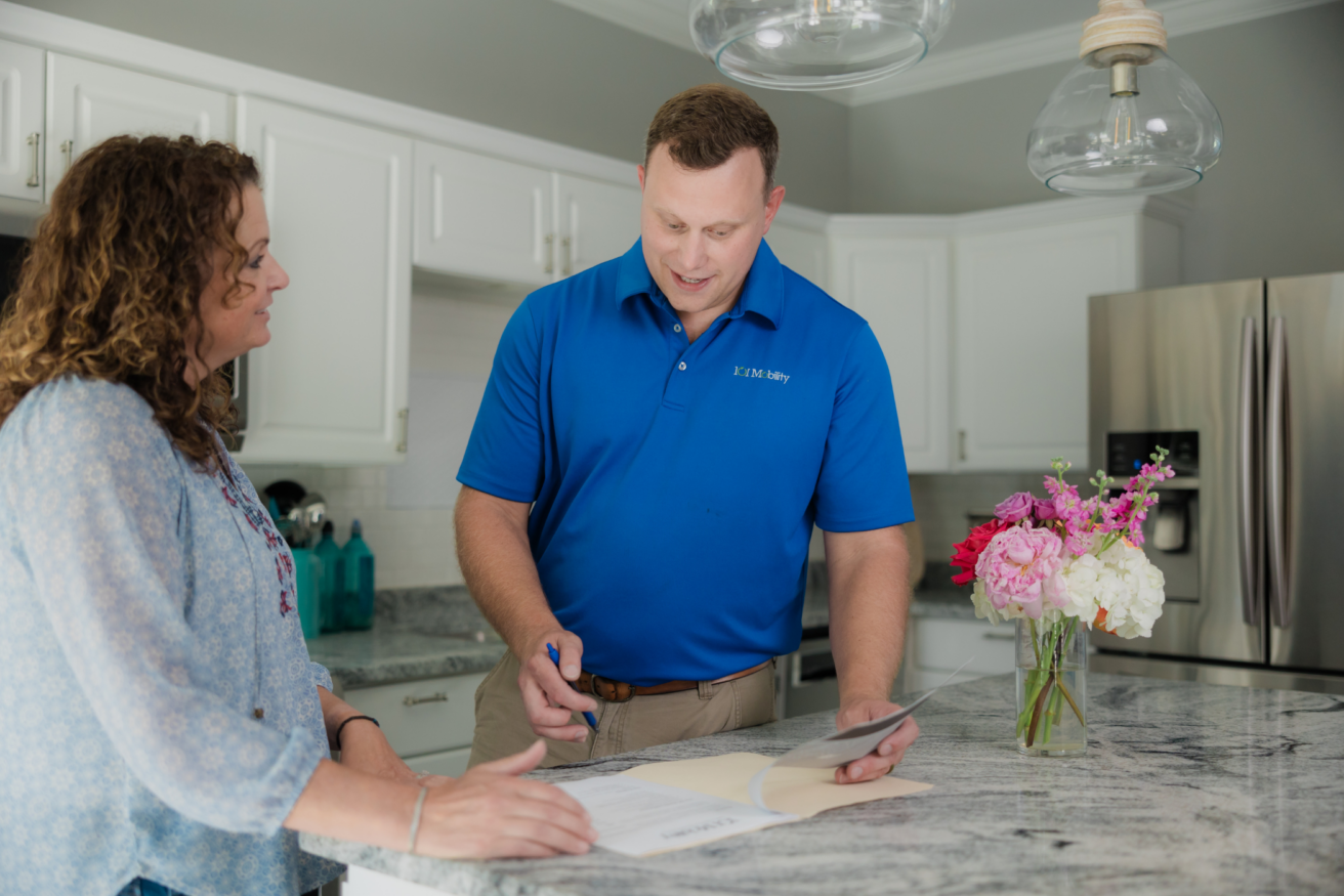 A man in a blue shirt is showing a piece of paper to a woman in a light-colored shirt while standing at a kitchen counter adorned with a vase of flowers.