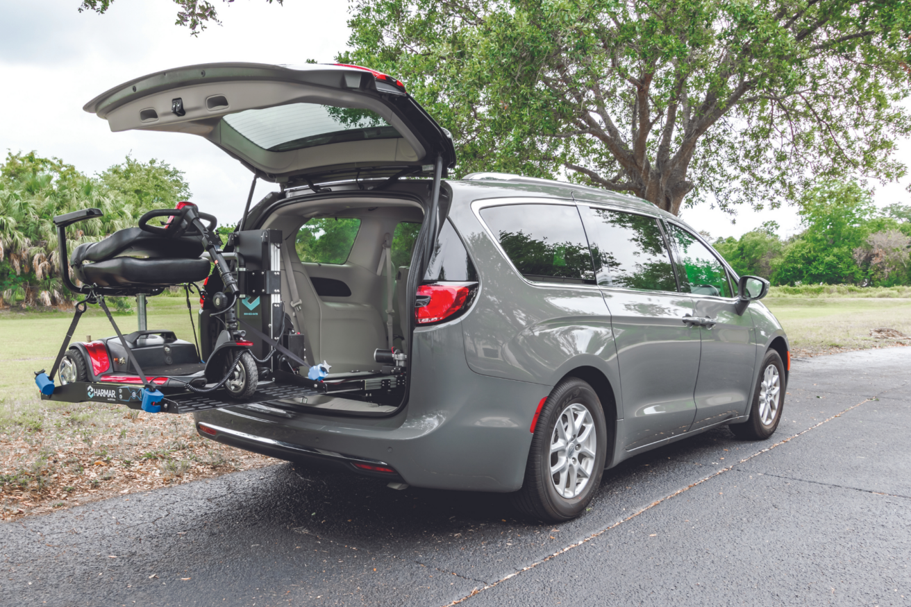 A gray minivan with an open rear hatch shows a mobility scooter hoist lifting a scooter into the vehicle. The van is parked on a paved surface with trees in the background.