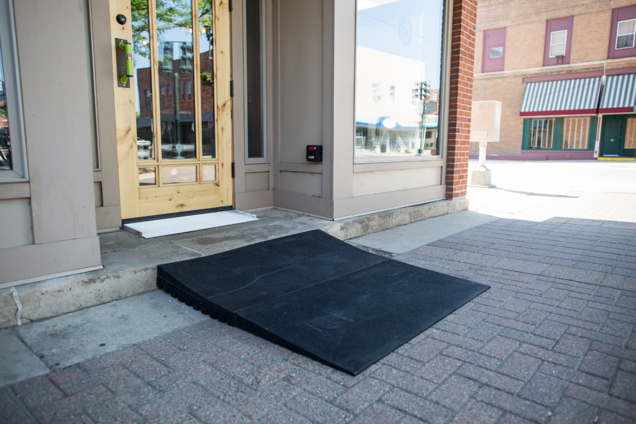 An accessible ramp leading to the entrance of a building, placed over a small step on a brick-paved sidewalk.