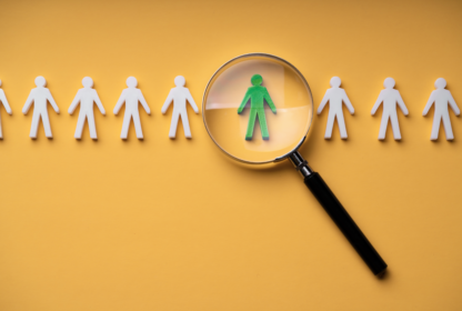 A magnifying glass highlights a green paper figure among a row of white paper figures on a yellow background.