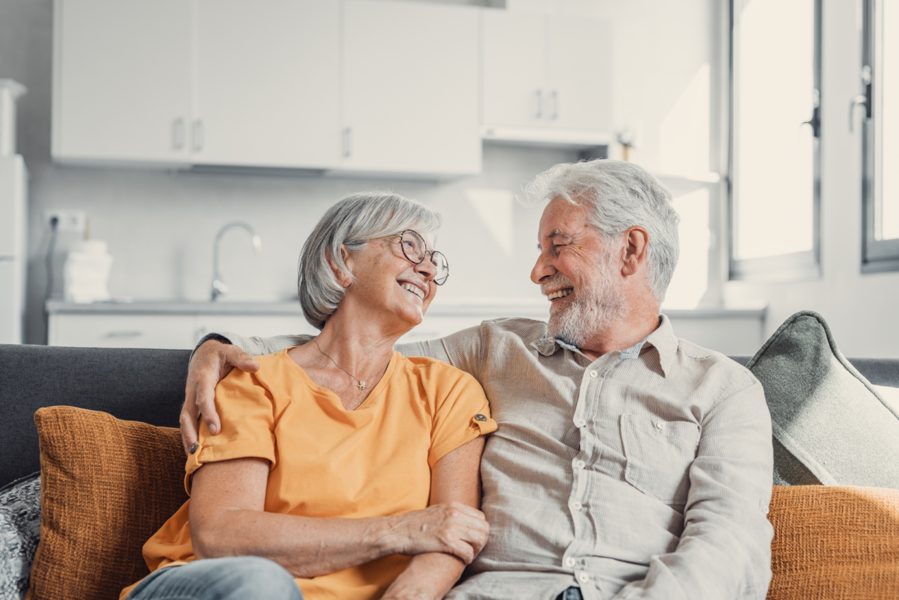 An elderly couple sits on a sofa, smiling and looking at each other, with the woman's arm around the man's shoulder in a bright living room.