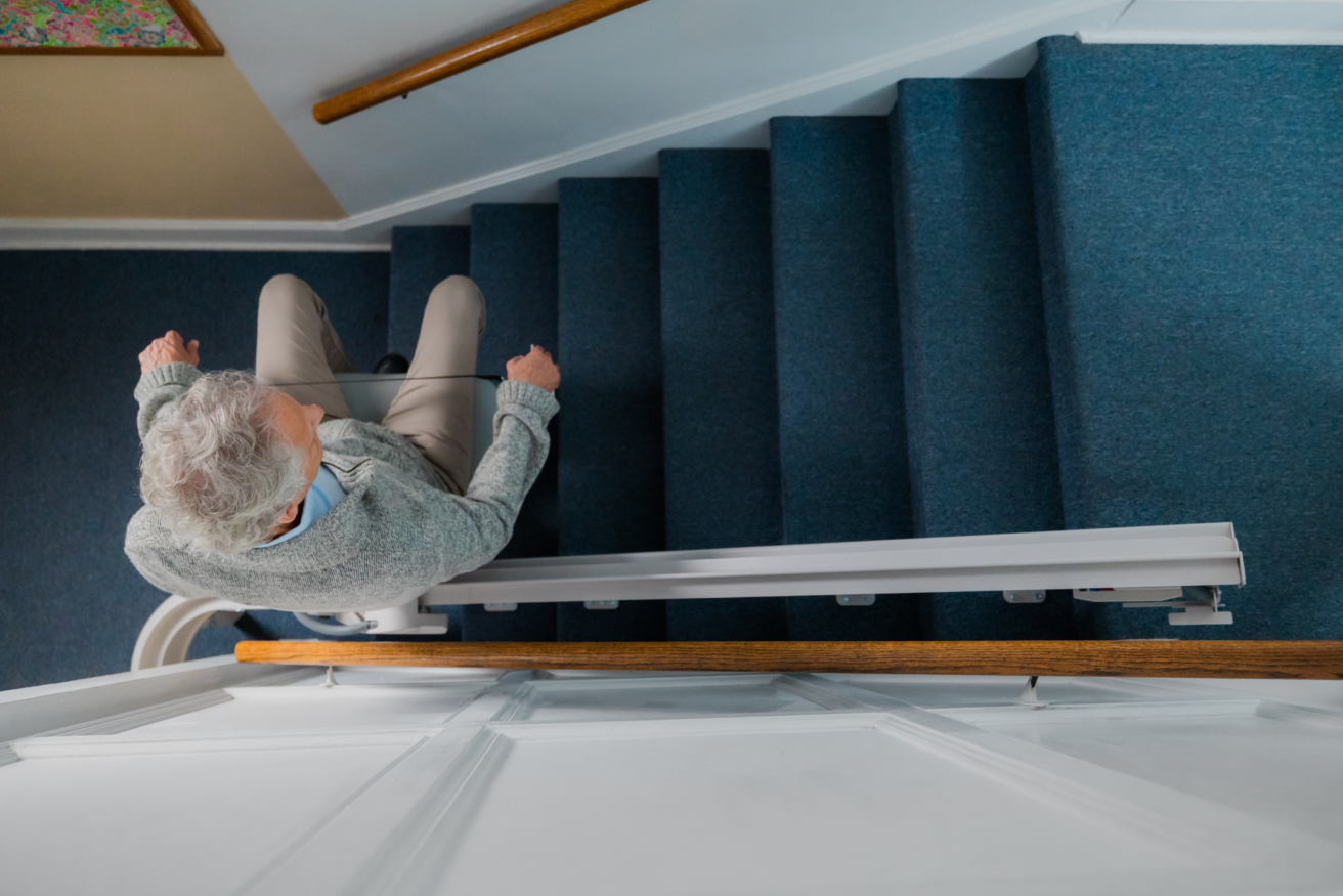 An older individual with gray hair rides a stairlift up a blue-carpeted stairway, viewed from above.