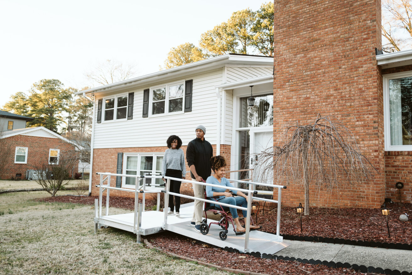 A family of three stands outdoors on a wheelchair ramp leading to the entrance of a house. The person in the wheelchair is being accompanied by two adults.