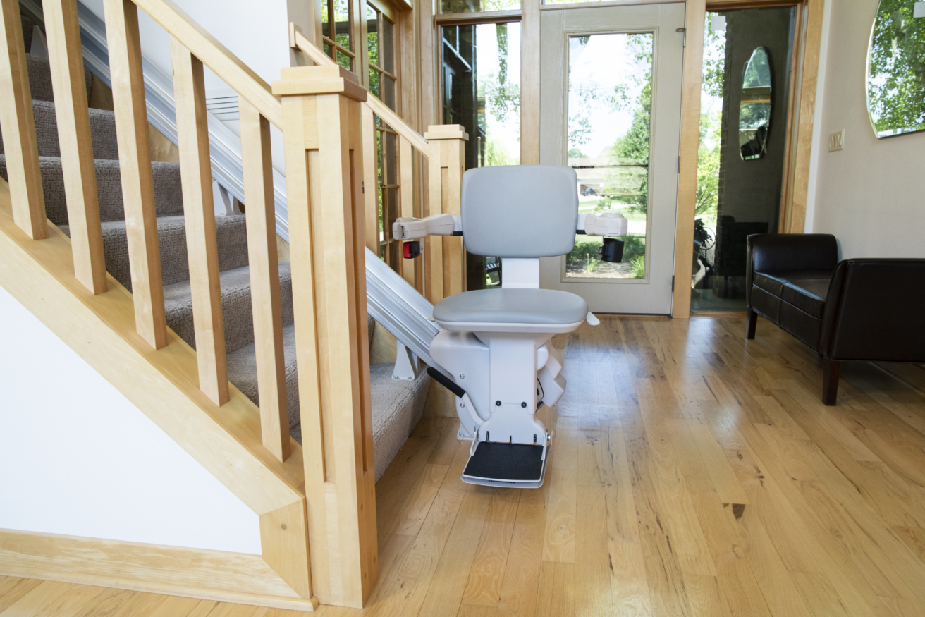 A home stairlift installed alongside a wooden staircase in a well-lit living space, with large windows in the background.