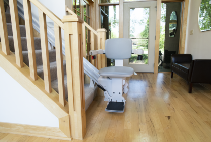 A home stairlift installed alongside a wooden staircase in a well-lit living space, with large windows in the background.