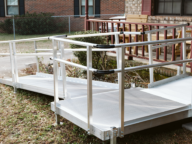 Aluminum wheelchair ramp leads to a porch with red railing, providing accessible entry to a brick house in a yard with grass and a chain-link fence.