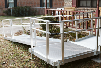 Aluminum wheelchair ramp leads to a porch with red railing, providing accessible entry to a brick house in a yard with grass and a chain-link fence.