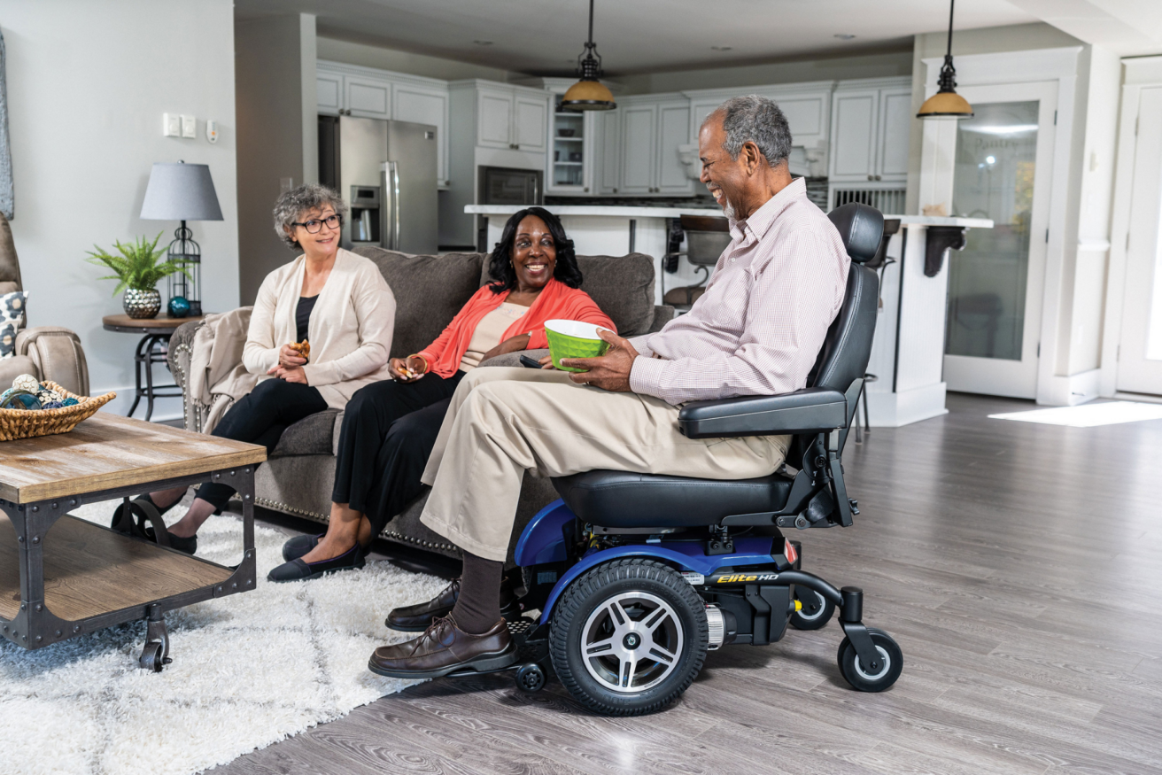 Three people are seated in a living room. One person is in a power wheelchair, while the other two are sitting on a sofa. The room is modern with a kitchen in the background.