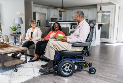 Three people are seated in a living room. One person is in a power wheelchair, while the other two are sitting on a sofa. The room is modern with a kitchen in the background.