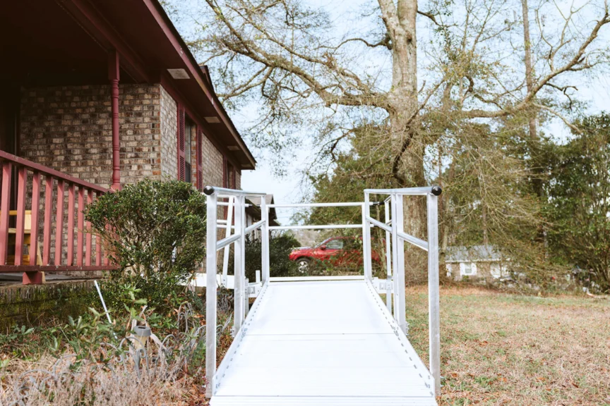 A metal accessibility ramp leads to the entrance of a brick house with a red roof, surrounded by trees and grass.