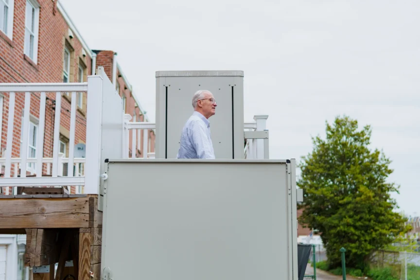 An elderly man stands on a residential outdoor lift near brick houses and a tree.