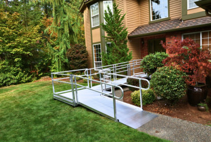 A house with a front yard features a metal accessibility ramp leading up to the entrance, surrounded by various trees and shrubs.