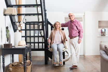 An elderly woman sits on a stairlift at the base of a staircase in a modern home, while an elderly man stands beside her, both smiling. Various household items are visible in the background.