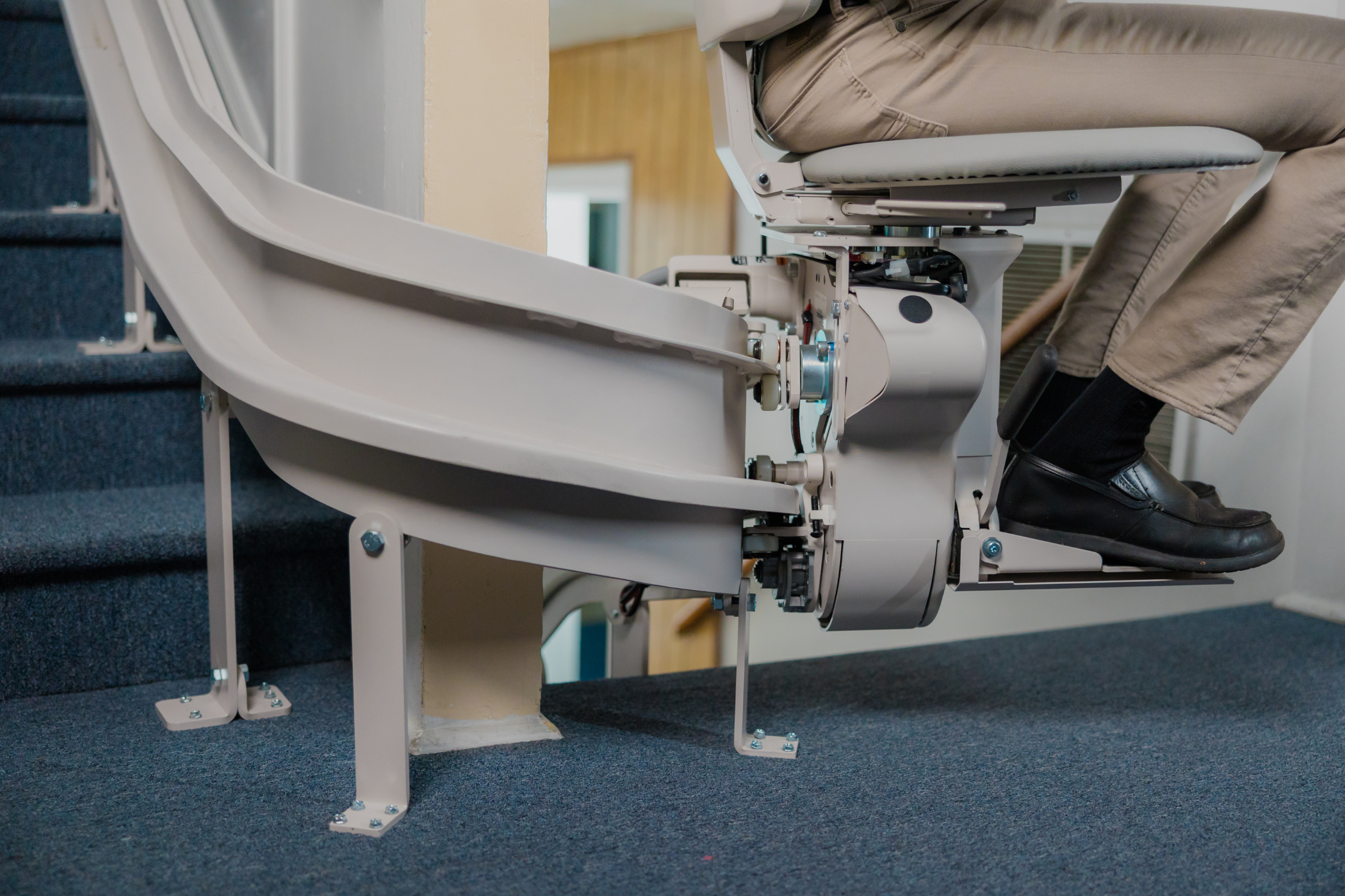 A person is seated on a stairlift chair attached to stairs, using it to ascend or descend. The stairlift mechanism and part of the user's lower body are visible.