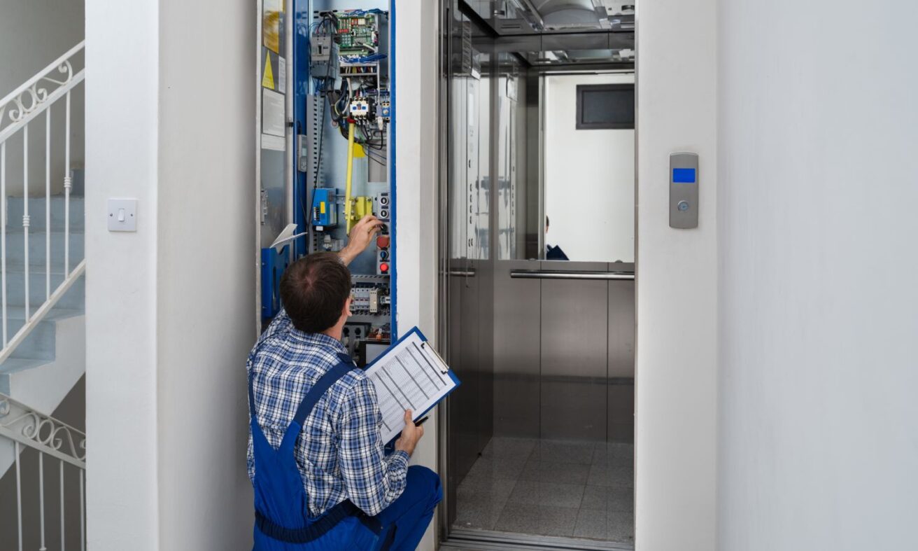 A technician in blue overalls works on the control panel of an elevator, holding a clipboard with a checklist.