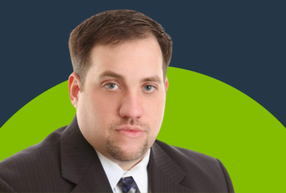 A man with short brown hair and a trimmed beard, wearing a dark suit and tie, is seen posing against a blue and green background.