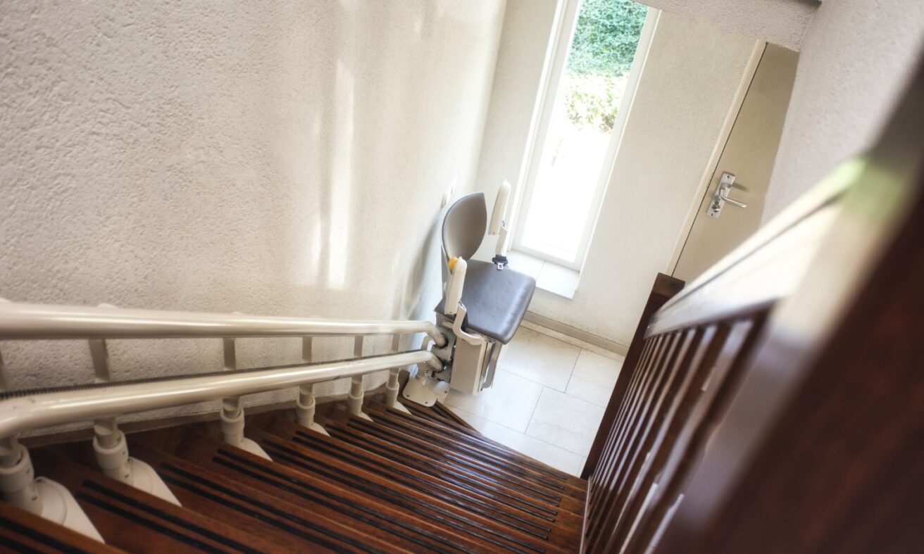 A stair lift is installed on a wooden staircase, leading down to a small, sunlit landing area with a door and window.