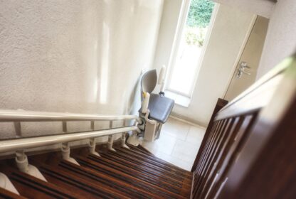 A stair lift is installed on a wooden staircase, leading down to a small, sunlit landing area with a door and window.