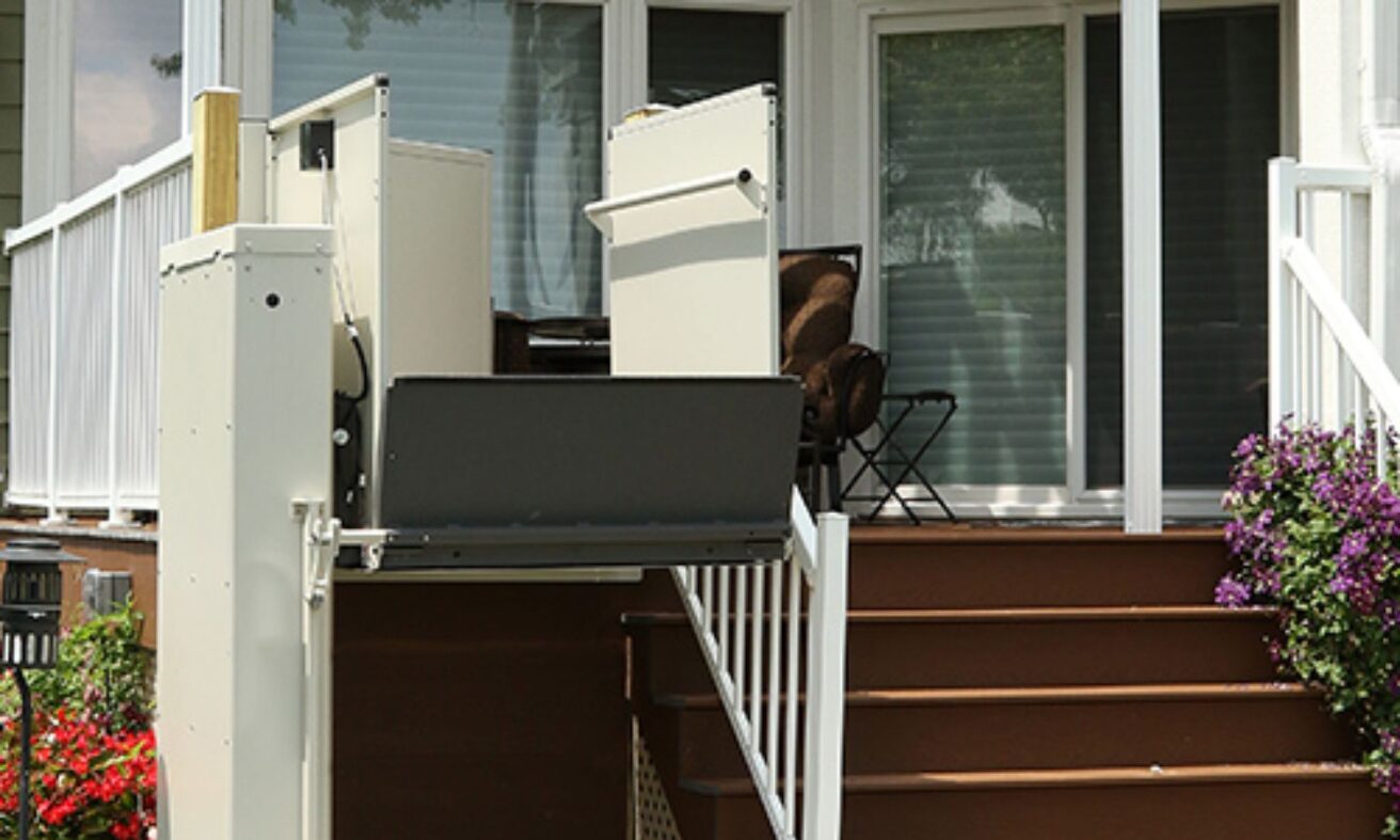 A residential wheelchair lift is installed adjacent to a set of stairs leading to a patio with sliding glass doors and white railings.