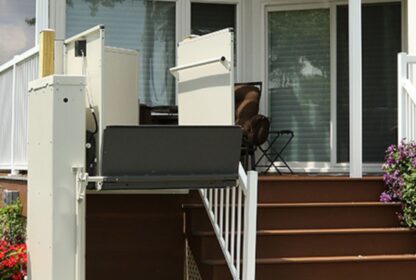 A residential wheelchair lift is installed adjacent to a set of stairs leading to a patio with sliding glass doors and white railings.