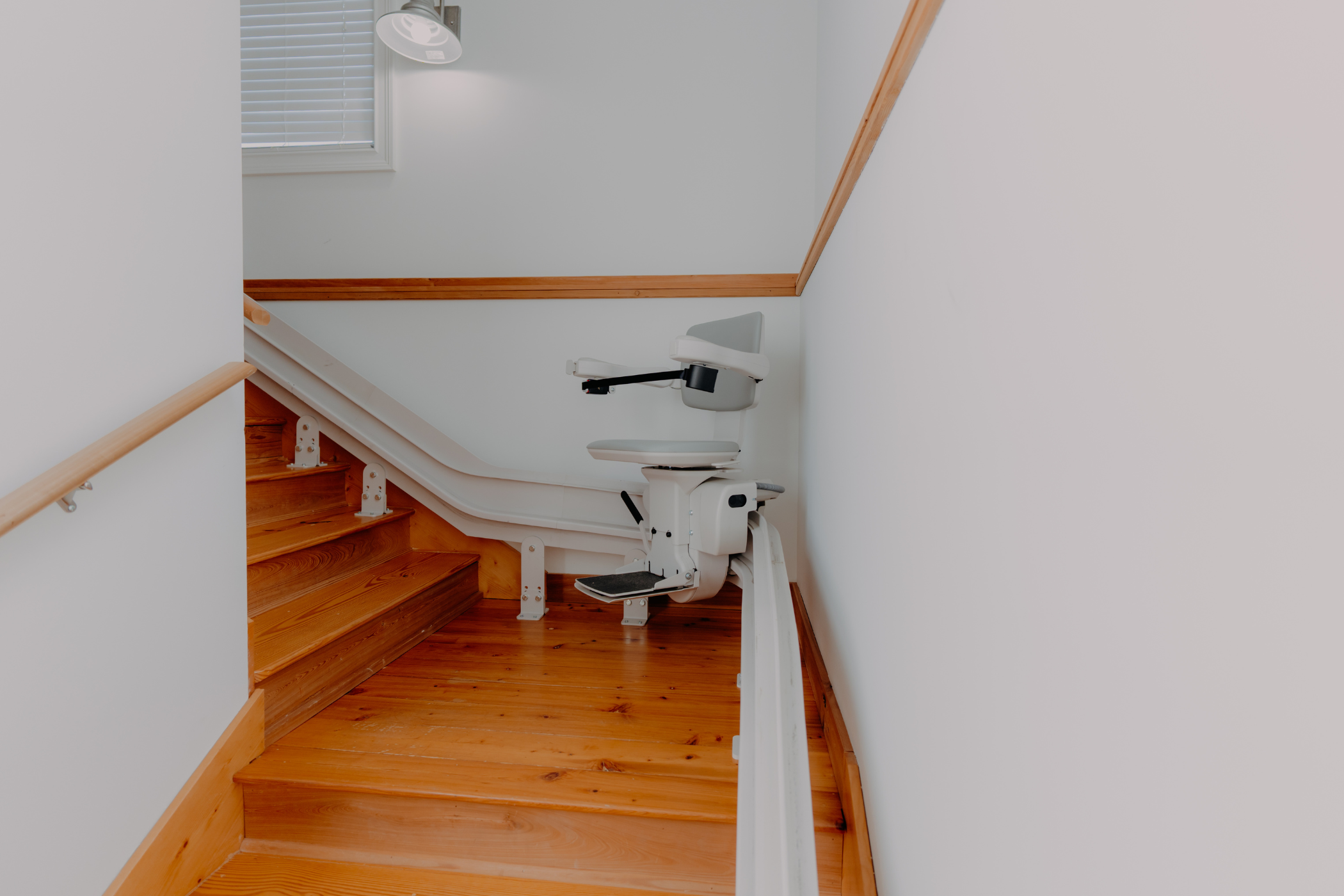 A stairlift is installed along a wooden staircase with white walls and a wooden handrail.