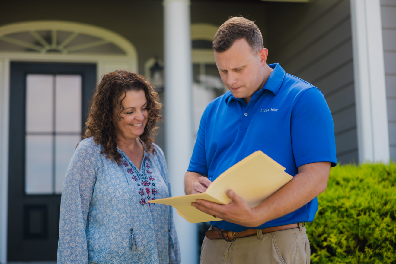 A man in a blue polo shirt shows a document to a woman standing outside a house.