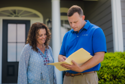 A man in a blue polo shirt shows a document to a woman standing outside a house.