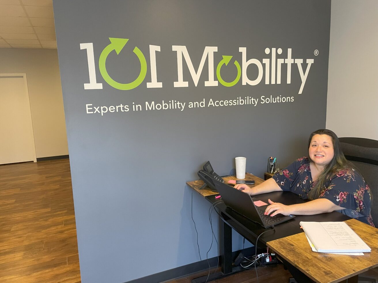 A woman works at a desk in an office with a sign on the wall that reads “101 Mobility: Experts in Mobility and Accessibility Solutions.”.