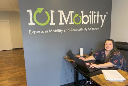 A woman works at a desk in an office with a sign on the wall that reads “101 Mobility: Experts in Mobility and Accessibility Solutions.”.